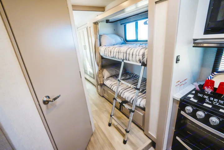 Compact RV rental interior featuring cozy bunk beds with a ladder, a bright window, and a functional kitchen area with an oven.. Thor Geneva 2026