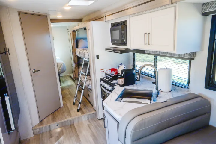 Inside an rv rental: functional kitchenette with stove and sink, bunk beds, and a separate bedroom beyond the door.. Thor Geneva 2026