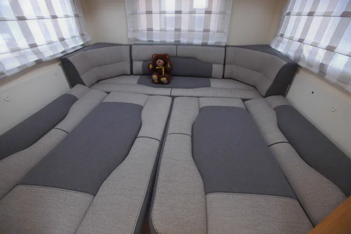 Cozy rv rental interior: Rear seating converted to a bed with two-tone grey cushions & a teddy bear. Striped window blinds.. rollerteam 747 2021