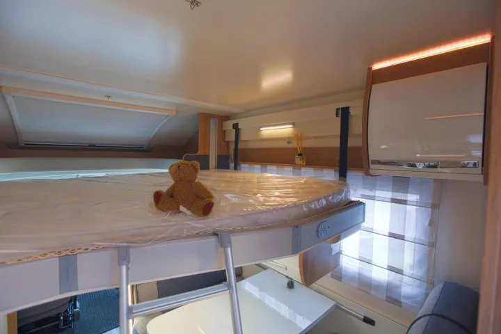 Cozy RV rental interior featuring an elevated bed with a teddy bear, an access ladder, and a compact dining area.. rollerteam 747 2021