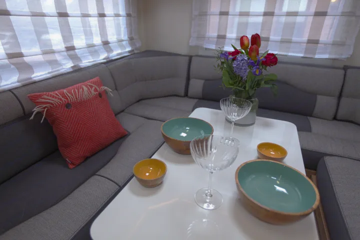 Inside a modern RV rental: a cozy dinette with U-shaped seating, a white table, colorful bowls, glasses, and fresh flowers.. rollerteam 747 2021