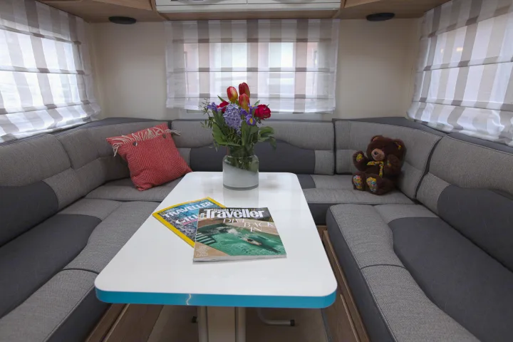 Comfortable RV rental dinette with U-shaped seating, white table, flowers, travel magazines, and a teddy bear.. rollerteam 747 2021