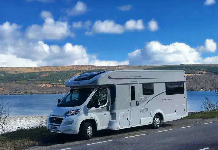 White RV rental parked by a scenic lake and hills under a blue sky. Perfect for an adventurous road trip exploring nature.. rollerteam 747 2021