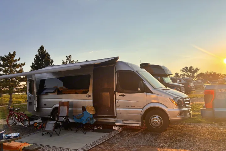 Relaxing by an RV rental camper van at sunset. Chairs, bike, and awning create a cozy campsite scene amidst trees and fields.. Mercedes-Benz Sprinter RV Motorhome Campervan 2017