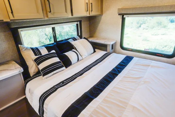Cozy RV rental bedroom featuring a striped bed, side tables, overhead cabinets, and scenic views from the windows.. Thor Geneva 2026