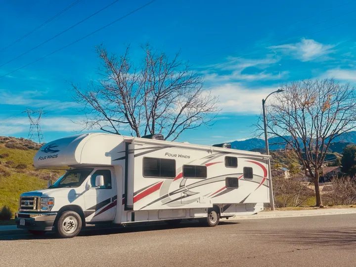 Four Winds Class C RV rental parked on a sunny road with hills, bare trees, and distant mountains under a blue sky.. Thor Four Winds 2022