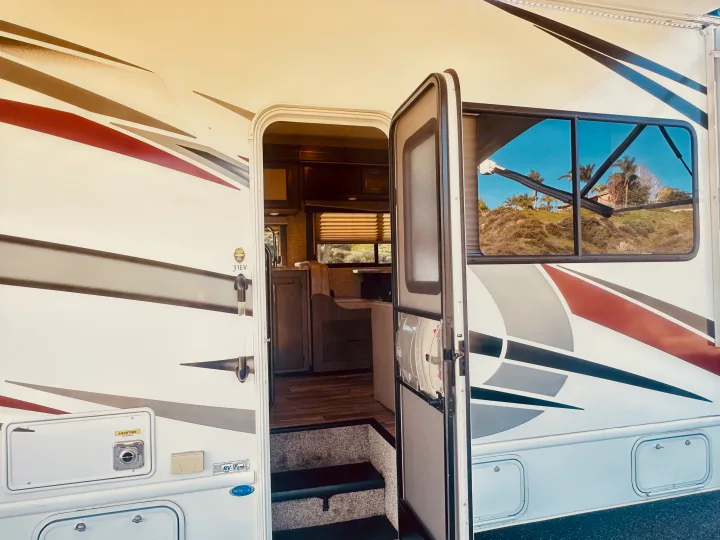An open door on this rv rental reveals a cozy interior, with a window reflecting a sunny hillside and palm trees outside.. Thor Four Winds 2022