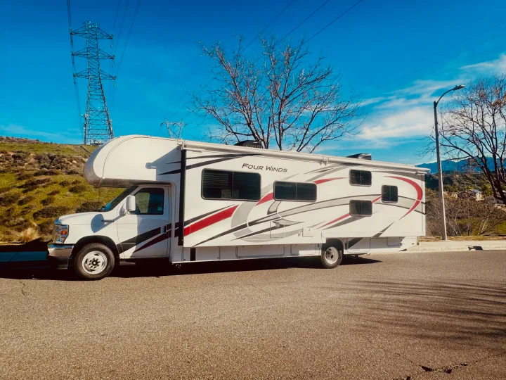 This white Four Winds Class C RV rental, parked on asphalt with hills and power lines in the background, awaits your adventure.. Thor Four Winds 2022
