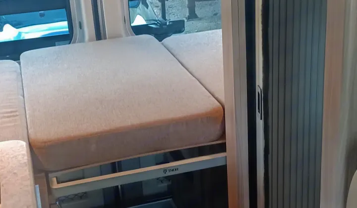 Comfortable light beige bed/seating in an RV rental, featuring a THOR frame, outdoor window view, and a rolling side door.. Thor Scope 2023