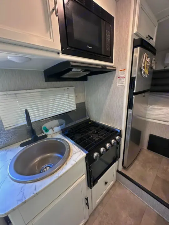 Inside a clean RV rental: a compact kitchen with a sink, gas stove, microwave, and stainless steel refrigerator.. Coachmen Crosstrek 23XG 2021
