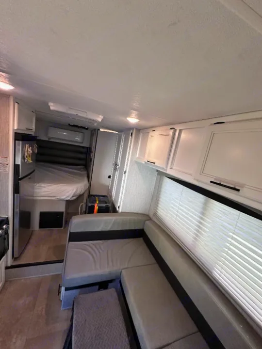 Interior of a modern rv rental, featuring an L-shaped dinette, cozy bed, kitchen area, and white overhead cabinets.. Coachmen Crosstrek 23XG 2021