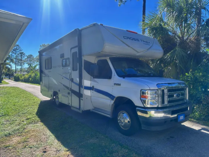 A white and blue Coachmen Cross Trek rv rental, parked on a sunny driveway, surrounded by palm trees and greenery.. Coachmen Crosstrek 23XG 2021