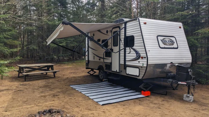 Our cozy RV rental parked at a wooded campsite, featuring an extended awning, picnic table, and fire pit for a relaxing stay.. Coachmen Viking 2017