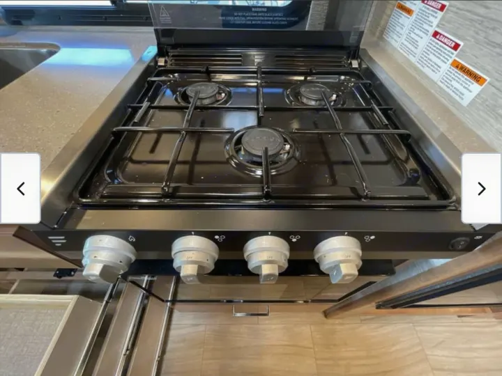 RV rental kitchen: A close-up of a 4-burner gas stovetop with control knobs, integrated into a counter. Safety warnings visible.. Thor Quantum 2021