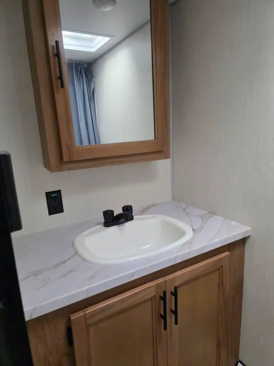 RV rental bathroom vanity: white sink, black faucet, marble-look counter, wood cabinet, mirror, & power outlet for convenience.. Forest River Cherokee Wolf Pup 2025