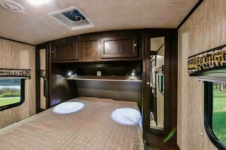 RV rental bedroom with overhead dark wood storage, cozy bed lit by bedside lamps, and a window to scenic views.. Cruiser RV Shadow Cruiser 2019