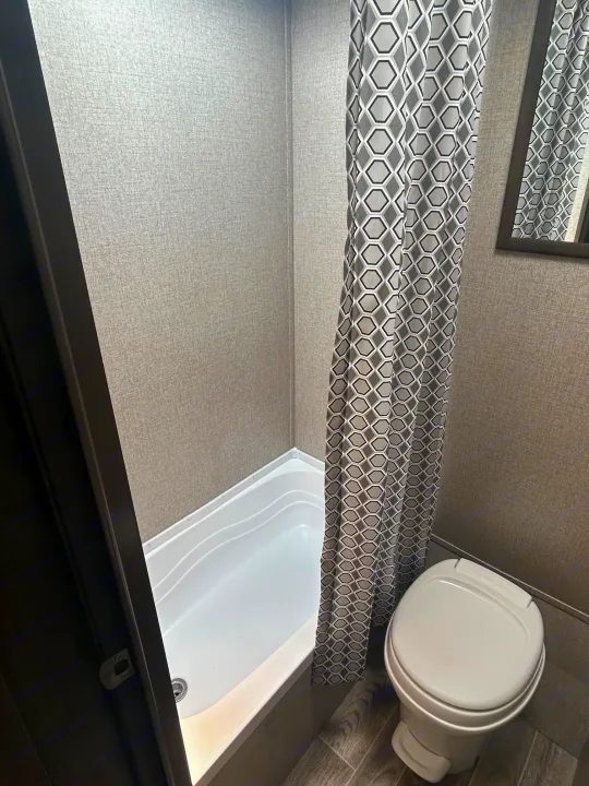 Compact RV rental bathroom features a white shower/tub, hexagon-patterned curtain, & toilet. Textured gray walls.. Jayco Jay Flight SLX 2021
