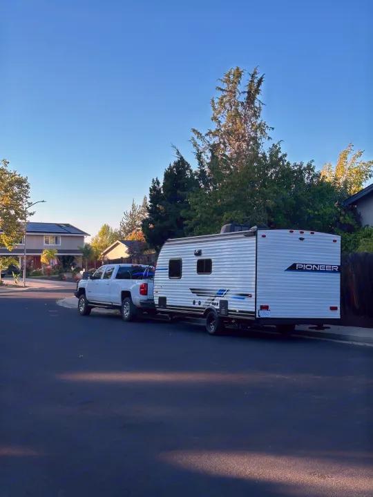 White pickup truck with a Pioneer RV rental travel trailer parked on a residential street under a clear blue sky.. Heartland RVs Pioneer 2025