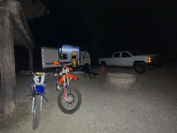 Nighttime RV rental camp: travel trailer, pickup, dirt bikes, and fire pit on gravel, lit by RV awning light.. Heartland RVs Pioneer 2025
