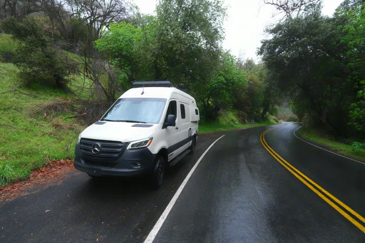 White Mercedes Sprinter rv rental camper van on a wet winding road. Ideal for scenic road trips and outdoor adventures.. Mercedes Winnebago 2024