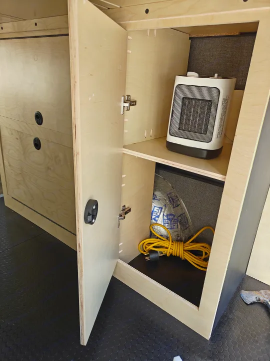 Inside this RV rental, a DIY plywood cabinet is open, revealing a portable heater, coiled yellow extension cord, and insulation for storage.. Mercedes-Benz Sprinter 2017