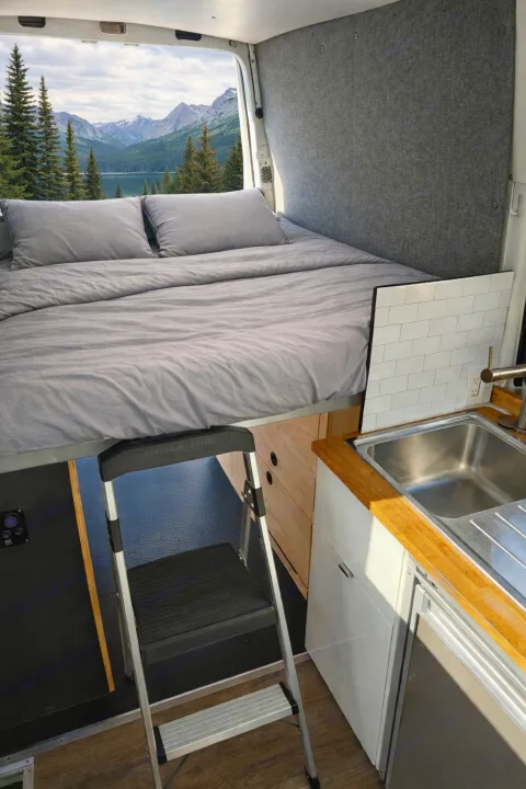 Cozy rv rental interior: elevated bed, kitchen with sink, and a step ladder. Outside, a stunning mountain lake view.. Mercedes-Benz Sprinter 2017