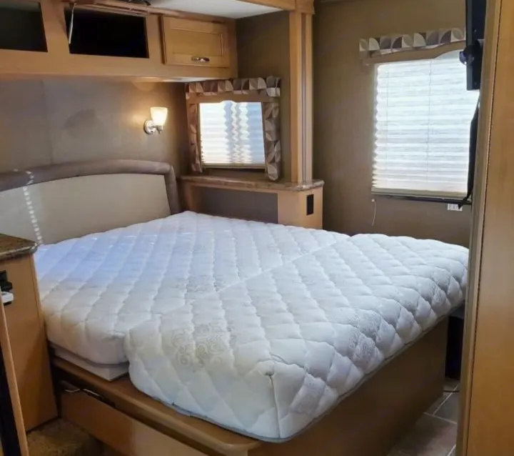 Experience comfort in this RV rental's bedroom, featuring a plush, white mattress, overhead storage, and a window with blinds.. Thor Chateau 2016