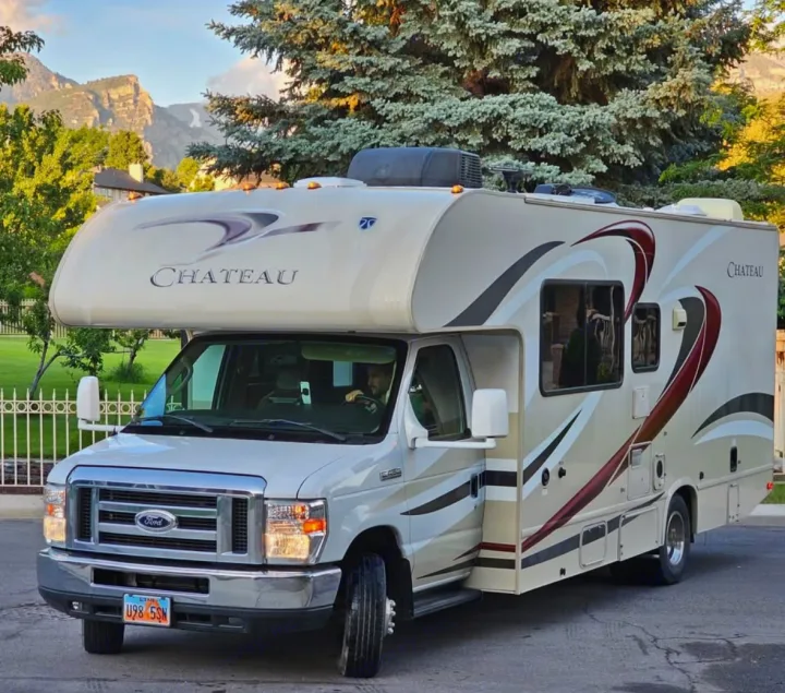 White Chateau Ford Class C RV rental with a driver, parked on a street. Mountains and trees in the scenic background.. Thor Chateau 2016