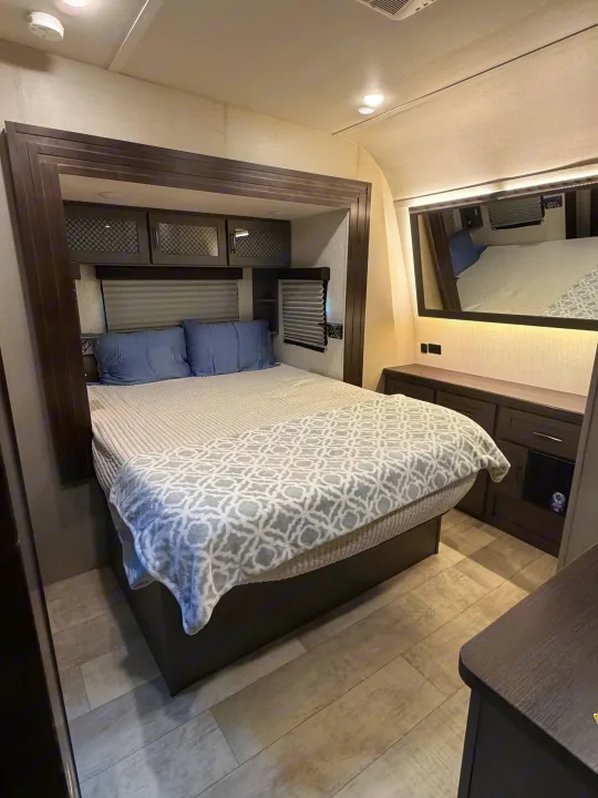 Cozy RV rental bedroom with a comfortable bed, dark wood furniture, light flooring, and a mirrored wall.. Grand Design Momentum Toy Hauler 2022