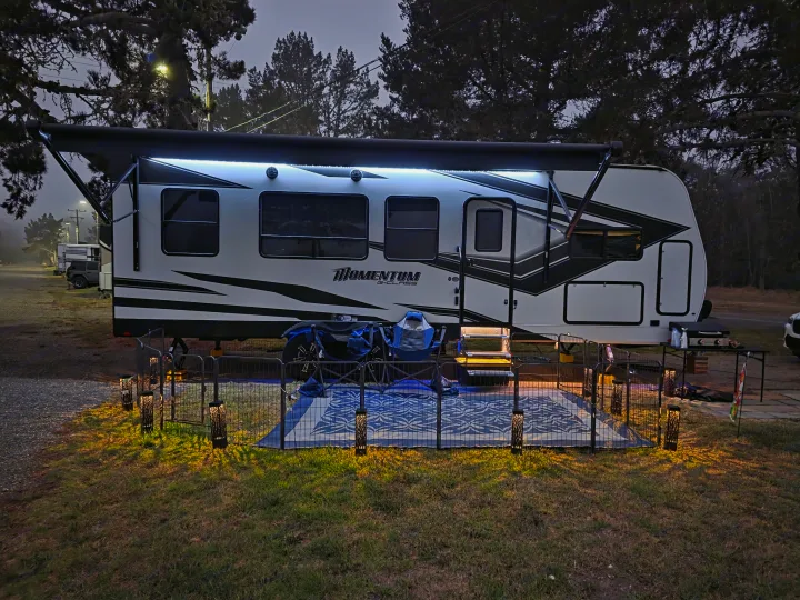 RV rental setup: A lit Momentum toy hauler with an extended awning, fenced patio, ATV, and grill at a cozy campsite by night.. Grand Design Momentum Toy Hauler 2022