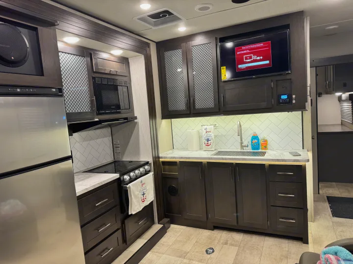 Inside an rv rental: a modern kitchen area with stainless steel fridge, microwave, stove, sink, and TV. Dark cabinets offer ample storage.. Grand Design Momentum Toy Hauler 2022