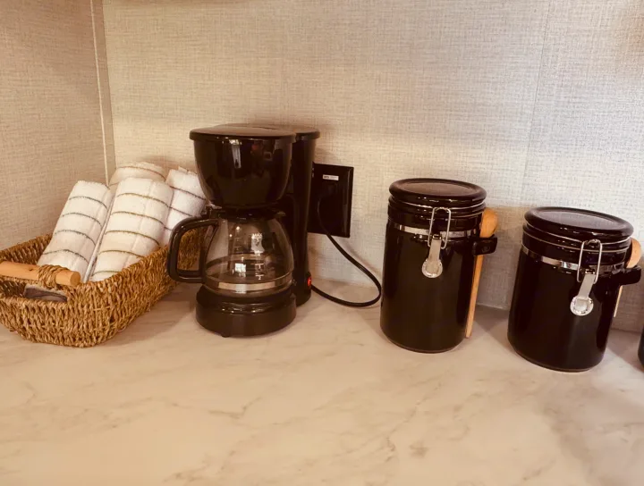RV rental's coffee station: black coffee maker, rolled towels in a basket, and two black canisters on the counter.. Grand Design Transcend One 2025
