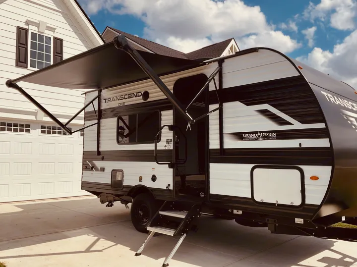 Grand Design Transcend One travel trailer, awning extended, parked in a sunny driveway by a white house. Ideal for RV rental!. Grand Design Transcend One 2025