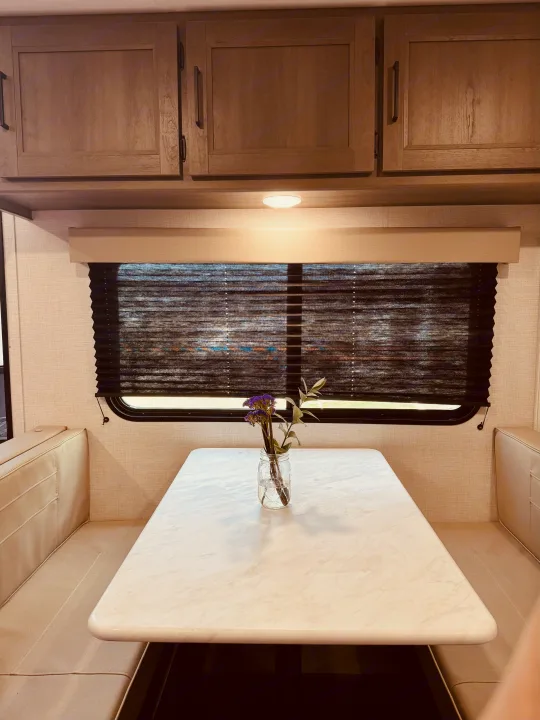 Inside an RV rental: a bright dinette area with light wood cabinets, beige upholstered benches, and a table holding a vase of flowers.. Grand Design Transcend One 2025