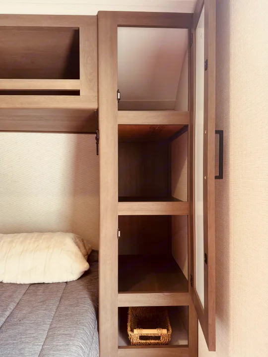 RV rental sleeping area featuring a bunk bed with soft bedding and an open storage cabinet with shelves, ready for your travel essentials.. Grand Design Transcend One 2025