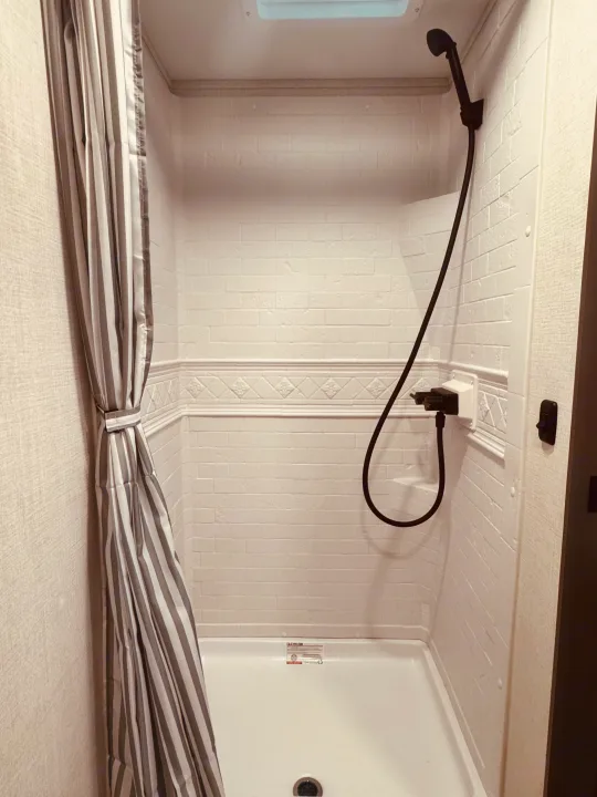 This bright, clean shower in an RV rental offers faux-brick walls, a striped curtain, and a black handheld shower fixture.. Grand Design Transcend One 2025