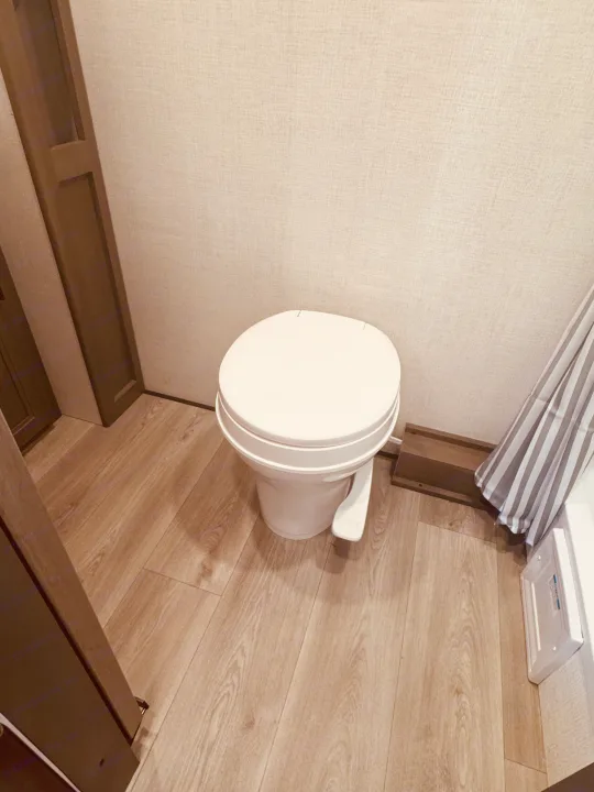 Compact RV rental bathroom features a white toilet with foot pedal, light wood-look floor, and textured beige walls.. Grand Design Transcend One 2025