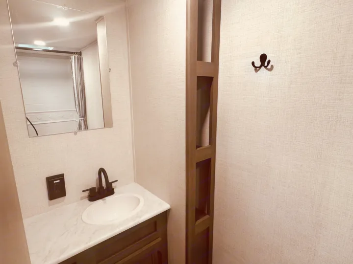 Compact RV rental bathroom: vanity with sink, mirror reflecting shower, built-in storage, and wall hook for convenience.. Grand Design Transcend One 2025