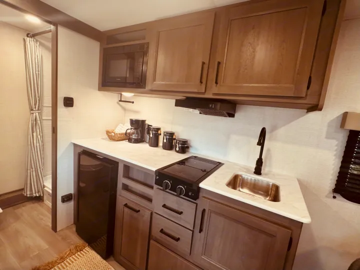 An rv rental galley with light wood cabinets, microwave, cooktop, sink, under-counter fridge, and a coffee station. Shower on left.. Grand Design Transcend One 2025