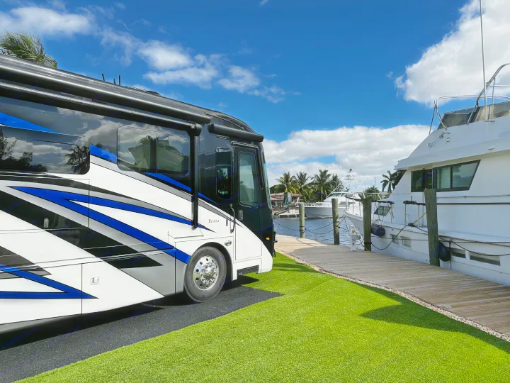 An RV rental with blue accents sits on green turf, overlooking yachts at a sunny waterfront dock with palm trees.. Entegra Coach Reatta 2021