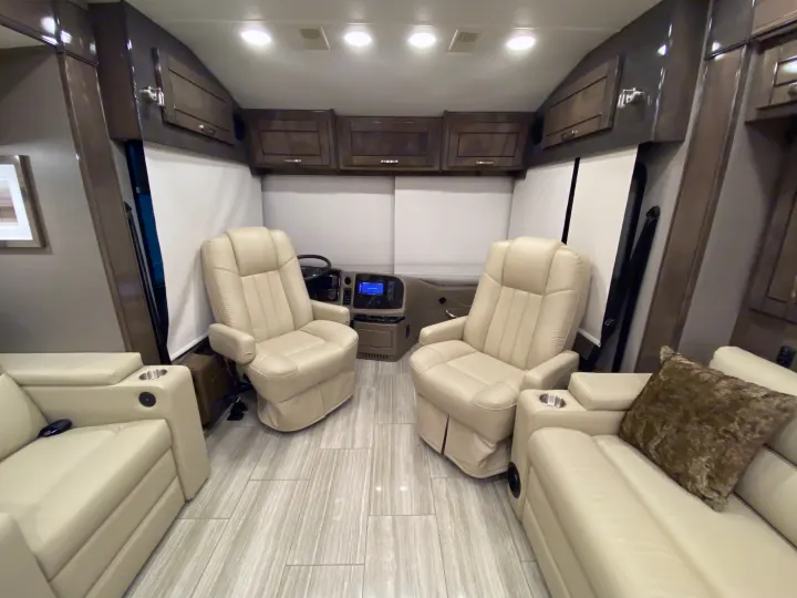 Modern rv rental interior: cream leather captain's chairs, lounge seating with cup holders, wood cabinets, and tiled floor.. Entegra Coach Reatta 2021