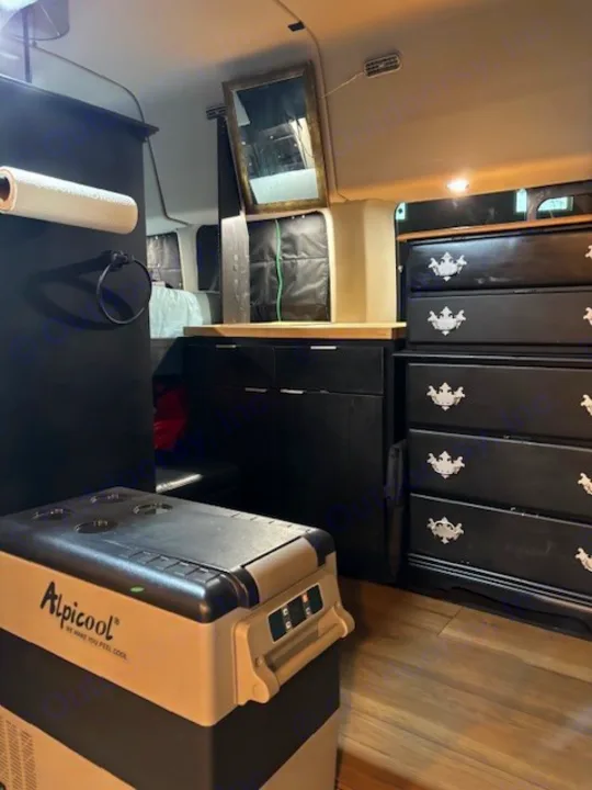 Inside this RV rental: Alpicool portable freezer, custom black cabinetry, and a dresser with decorative pulls, offering storage.. Ford Transit 2019