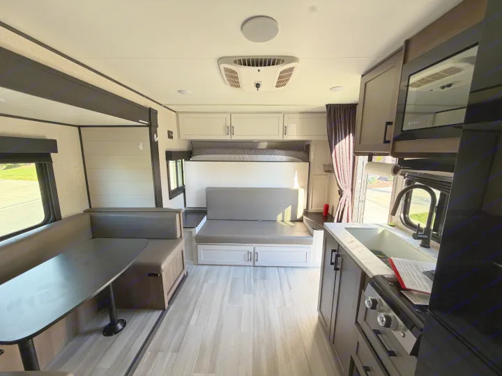 RV rental interior: dining booth, fold-down bed above bench, kitchen with sink/stove, overhead cabinets, and light wood floor.. Autumn Ridge 188BHS 2025