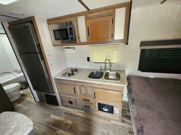 Bright rv rental kitchen with microwave, stove, sink, and fridge. Features wood-look flooring, storage, and adjacent bed/toilet area.. Gulf Stream Ameri-Lite Super Lite 2022