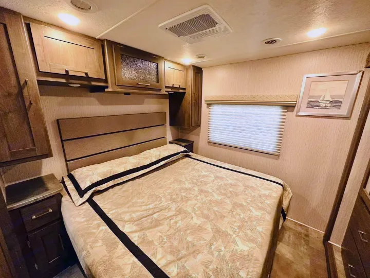 Comfortable RV rental bedroom with a tan patterned bed, upholstered headboard, wood cabinets, and window with blinds.. Coachmen Pursuit 31BH 2023