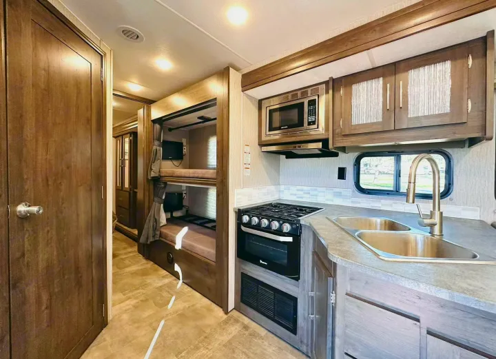 Inside an RV rental: a modern kitchen with stove, microwave, double sink, and a cozy bunk bed area with individual TVs.. Coachmen Pursuit 31BH 2023