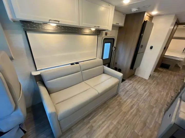 This bright RV rental interior offers a comfortable light-colored sofa, window shade, and modern wood-look flooring leading to a bedroom.. Thor Freedom Traveler 2025