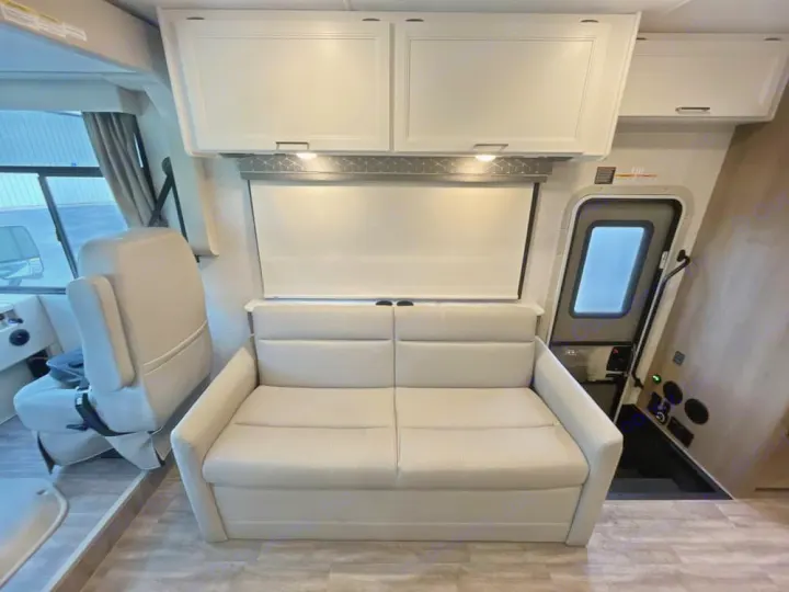 Bright RV rental interior features a light sofa, overhead storage, driver's seat, and entrance door.. Thor Freedom Traveler 2025