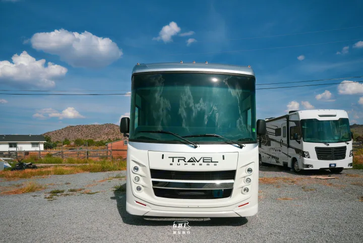 Front view of a white Travel Supreme RV for rv rental, parked on gravel. Another RV, a house, and hills in the background.. Jayco Travel Supreme A Series 27A 2025