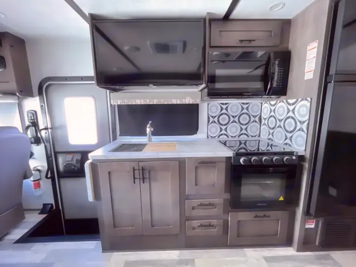 This modern RV rental kitchen interior features a stove, microwave, sink, and fridge. An exit door and TV are also visible.. FR3 FR3 Motorhome 2024
